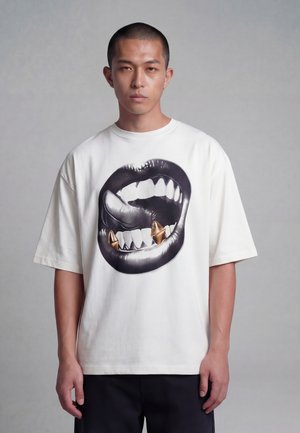 Giovane uomo che indossa una T-shirt bianca oversize con una grande grafica di labbra nere e borchie dorate a forma di zanne sul davanti.