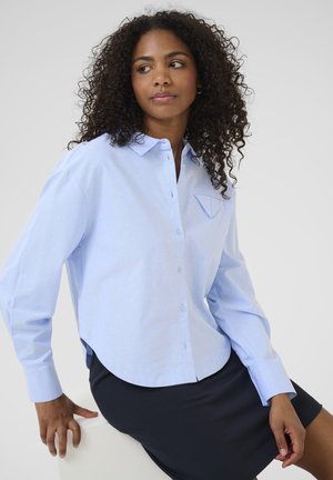 Light blue button-up shirt with a curved hem, long sleeves, and a chest pocket detail, made of smooth fabric. Worn with a dark skirt.