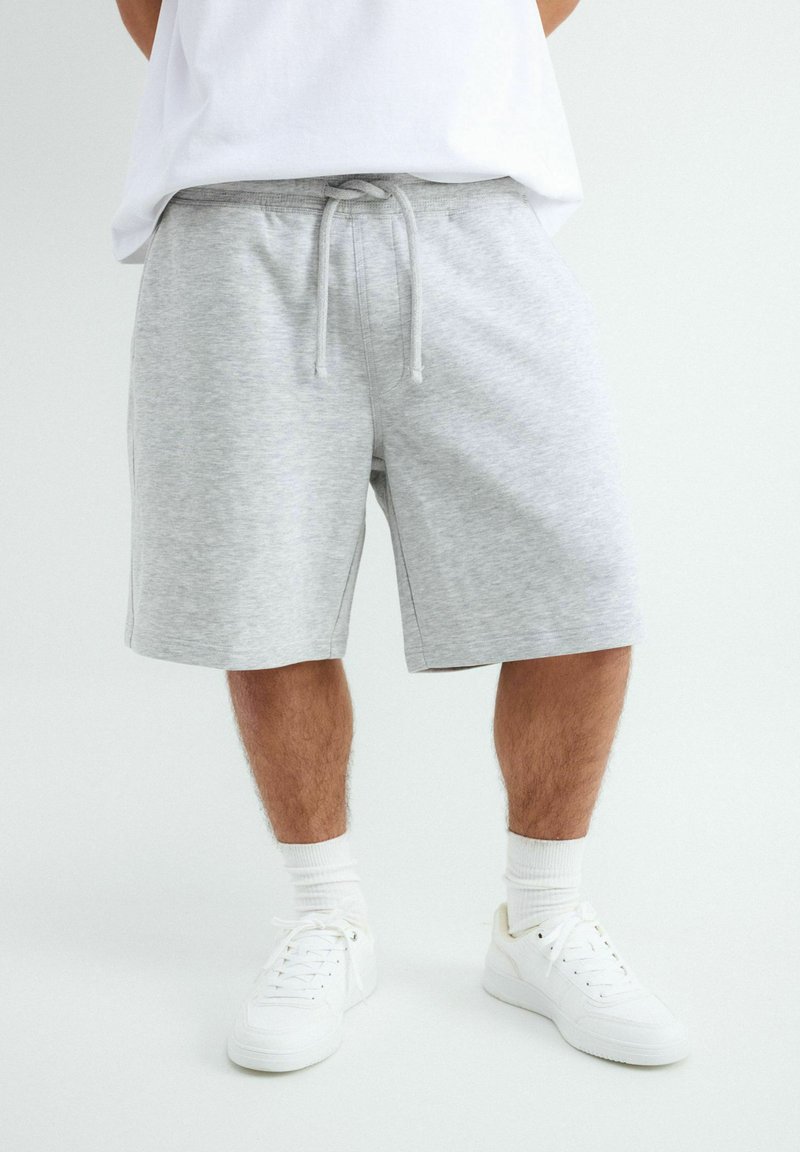 Homme portant un short gris clair à cordon, des chaussettes blanches et des baskets blanches, debout devant un fond clair uni.