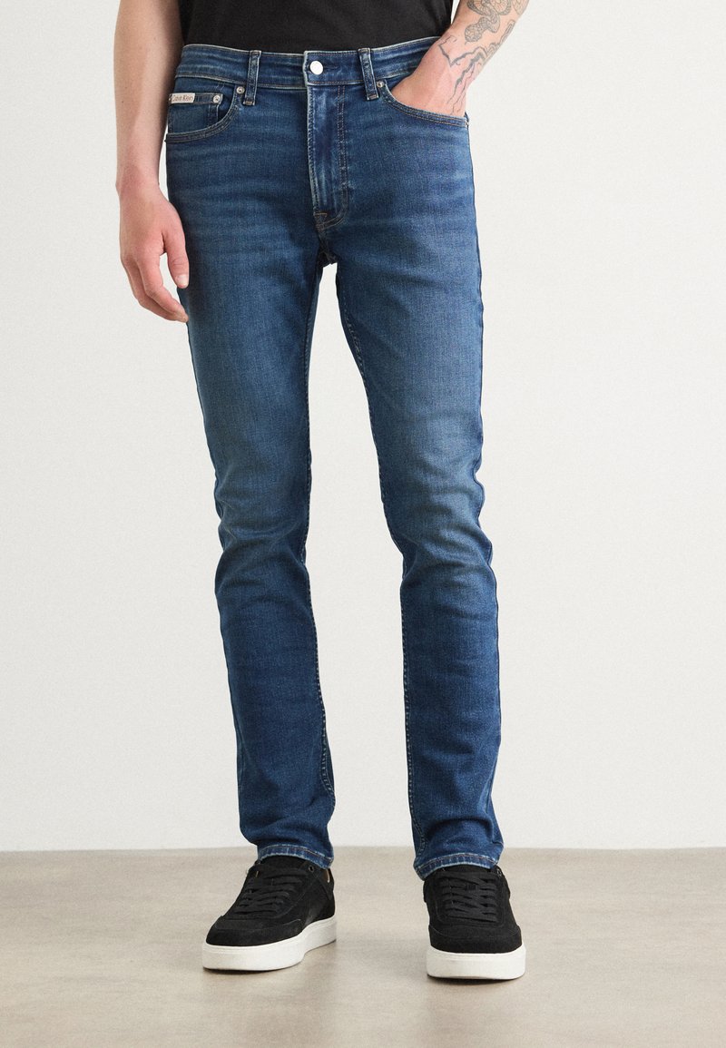 Dunkelblaue Slim-Fit-Jeans mit leichter Ausbleichung, vorderen Taschen und klassischem Bund. Kombiniert mit schwarzen Sneakern und einem schwarzen T-Shirt.