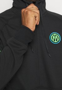 Haut à capuche zippé noir en tissu lisse, avec des poignets côtelés, une capuche avec cordon de serrage, et un logo circulaire avec des accents bleus et verts.