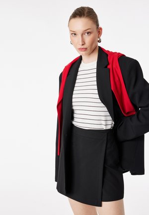 Kvinde iført sort blazer med rød foring over hvidt, stribet skjorte og sort nederdel, stående med hånden på hoften foran en hvid baggrund.