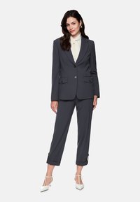 Dark gray tailored suit with two front pockets, single button closure, and rolled cuffs. Lightweight fabric, paired with a light-colored blouse and white heels.