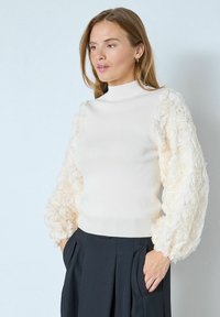 Kreemikas keha järgnev kõrge kaelusega kampsun, millel on tekstuurse kangaga kolme mõõtmelised lillemotiivid mahukatel varrukatel. Mustad püksid.