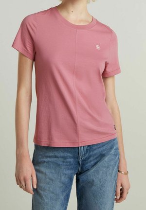 Kobieta w dopasowanym, jasnoróżowym, krótko rękawowym t-shircie w połączeniu z niebieskimi dżinsami, z minimalną biżuterią na lewej dłoni i nadgarstku.