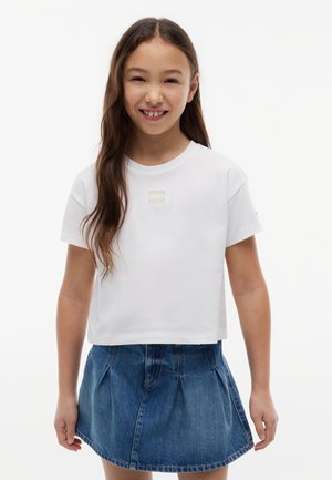 Ung pige med langt brunt hår smiler, iført en hvid kortærmet top og en blå denimnederdel, stående mod en hvid baggrund.