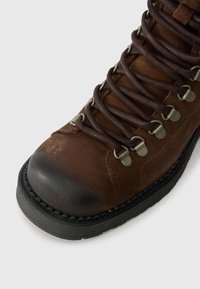 Ciemnobrązowy skórzany but trekkingowy z zaokrąglonym czubkiem, ciemną gumową podeszwą, metalowym systemem sznurowania oraz textured zamszowym wierzchem.