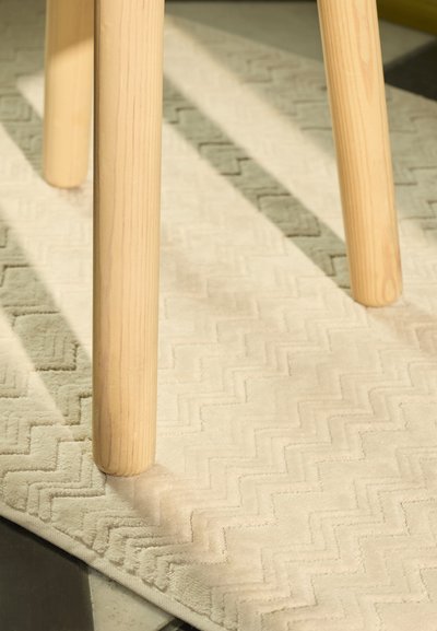 Perna de cadeira de madeira sobre um tapete texturizado de cor clara com um padrão em ziguezague, onde as fibras macias conferem uma aparência aveludada.
