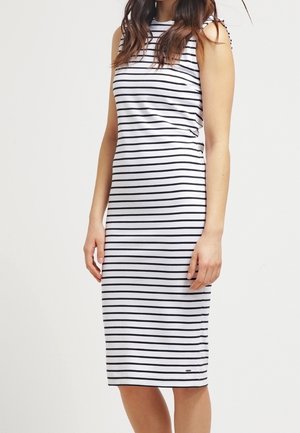 Vrouw met een mouwloze witte jurk tot aan de knieën met horizontale zwarte strepen, staand tegen een effen lichte achtergrond.