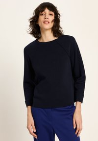 Frau mit schulterlangen dunklen Haaren, die ein schwarzes Langarmshirt und blaue Hosen trägt, steht vor einem schlichten hellen Hintergrund.