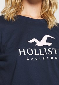 Camiseta de algodón color azul marino con un logo de gaviota rosa y texto blanco que dice "HOLLISTER CALIFORNIA" debajo del gráfico.