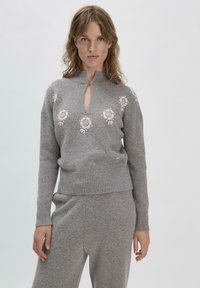 Grauer Strickpullover mit hohem Kragen, der mit rosafarbener Blumenstickerei über der Brust und einem Schlüssel-Loch-Design auf der Vorderseite versehen ist.
