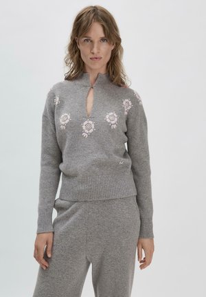 Grauer Strickpullover mit hohem Kragen, der mit rosafarbener Blumenstickerei über der Brust und einem Schlüssel-Loch-Design auf der Vorderseite versehen ist.