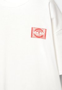 Bijela majica kratkih rukava s crvenim printanim oznakama na lijevoj strani prsiju, ukrašena dekorativnim elementima i tekstom.