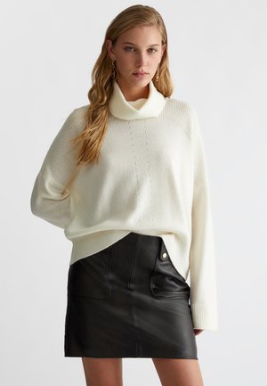 Maglione a collo alto color crema con maniche texturizzate, abbinato a una minigonna nera in pelle aderente con tasche anteriori e bottone a scatto.