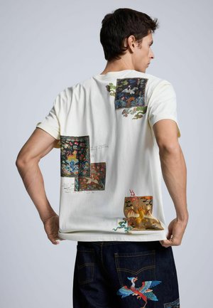 Uomo di spalle che indossa una camicia bianca e jeans scuri con disegni ricamati colorati di uccello e drago sulla schiena.