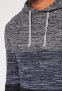 Man met een gebreide hoodie met horizontale grijze en marineblauwe strepen en grijze trekkoorden, die de bovenste torso en nek toont.