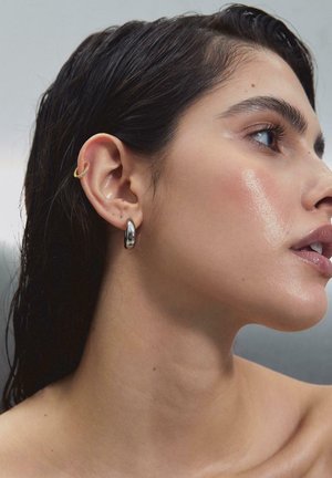 Profil af en kvinde med vådt mørkt hår, iført sølv øreringe og små gyldne øreklemmer på hendes venstre øre, med glat og strålende hud.