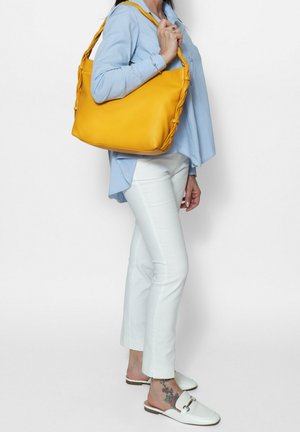 Sac à épaule en cuir jaune avec des détails tressés, texture lisse et design spacieux. Porté sur l'épaule contre une chemise bleu clair.