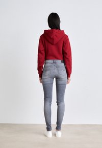 Rode hoodie, grijze skinny jeans, witte sneakers. Gladde texturen, casual pasvorm, zichtbare stiksels en een klein logo-label op de jeans.