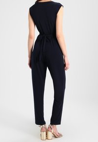 Combinaison bleu marine avec un corsage ajusté, des manches courtes et un détail de ceinture nouée à la taille. Le tissu est lisse, associé à des sandales à talons métalliques.