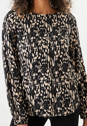 Frau trägt eine langärmelige Bluse mit einem schwarzen und beigen abstrakten gefleckten Muster, kombiniert mit schwarzen Hosen.