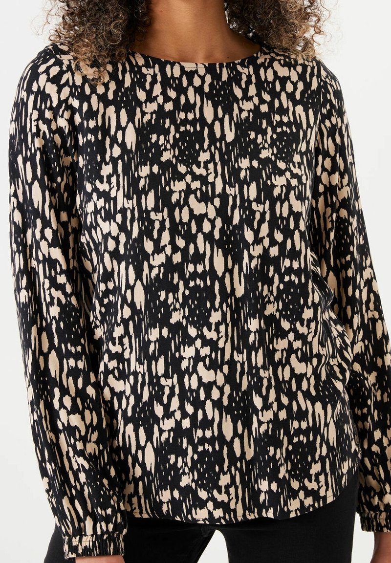 Femme portant un chemisier à manches longues avec un motif tacheté abstrait noir et beige, associé à un pantalon noir.