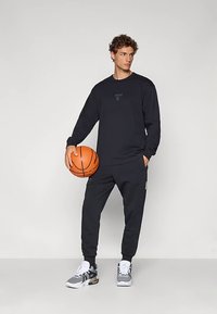 Μαύρη αθλητική μπλούζα με μακριά μανίκια που διαθέτει ένα μικρό λογότυπο. Συνδυάζεται με μαύρα παντελόνια jogging και γκρι αθλητικά παπούτσια, κρατώντας μια μπασκέτα.