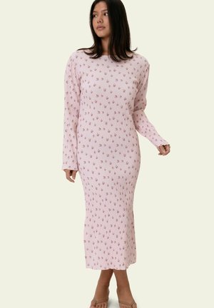 Vrouw draagt een lichtroze midi-jurk met lange mouwen en ribstructuur, voorzien van een klein bloemenpatroon, staande tegen een effen achtergrond.