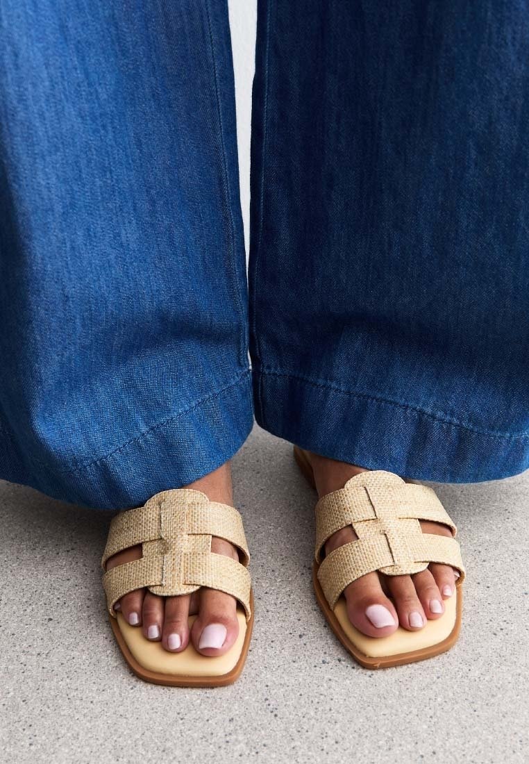 Personne portant un jean bleu à jambes larges et des sandales tissées beiges avec les ongles de pieds peints, debout sur un sol gris clair.