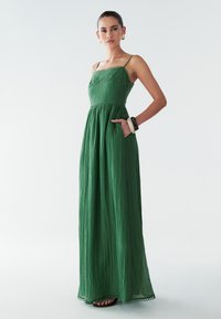 Zaļa maksi kleita ar spageti siksnām, strukturētu audumu un savilktu jostu. Ietver kabatas un viļņotu apakšmalu.