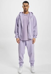 Hoodie lilas et jogging assorti en tissu doux, avec fermeture éclair, poches avant et chevilles resserrées, accompagnés de baskets blanches.