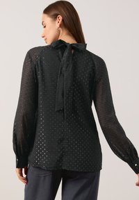 Schwarze Bluse mit strukturiertem Muster, durchsichtigen langen Ärmeln und einer Bindung am Rücken der Halslinie. Hat einen lockeren Schnitt und knöpfbare Manschetten.