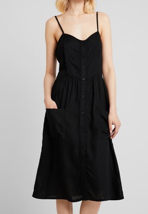 Woman wearing a black button-front midi dress with thin straps and two large front pockets, standing against a plain light background.