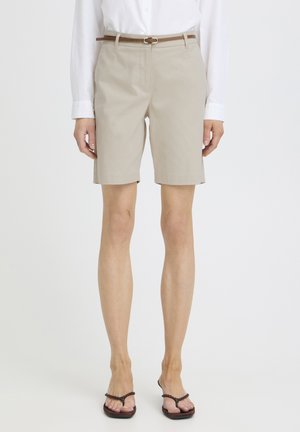 Kvinde iført beige knælange shorts med brunt bælte, hvid skjorte med lange ærmer og brune thongsandaler, stående mod en ensfarvet baggrund.