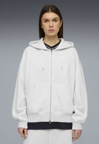 Νεαρή γυναίκα που φορά γκρι ανοιχτό φούτερ με φερμουάρ και εμπρόσθιες τσέπες, με λευκό λογότυπο Puma στο στήθος, στέκεται μπροστά σε φόντο γκρι διαβάθμισης.