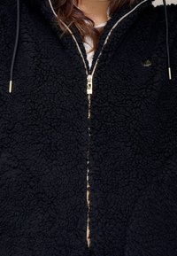 Chaqueta de forro polar negra con una superficie texturizada, cremallera dorada y cordones ajustables, que presenta un pequeño logo en la parte superior izquierda.