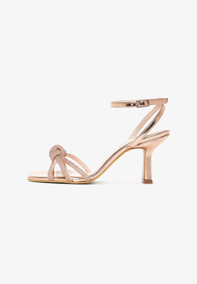 High-heeled sandal in rose gold with a glossy finish, featuring a knotted design, narrow straps, and a slim ankle strap for secure fit.