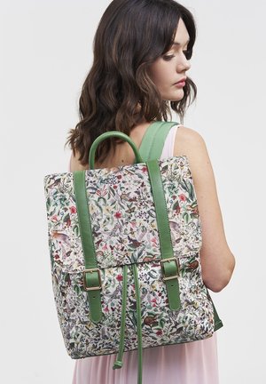 Mujer de cabello oscuro que lleva un vestido sin mangas y una mochila con estampado floral, con correas y hebillas verdes, en la espalda.
