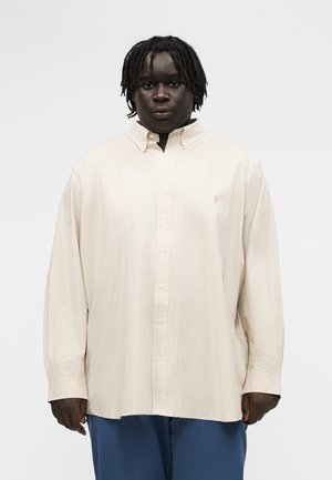 Άτομο με σκούρα πλεγμένα μαλλιά που φοράει μια φαρδιά μπεζ καρώ πουκάμισο με κουμπιά και μπλε παντελόνι, στέκεται μπροστά σε μια απλή φόντο.