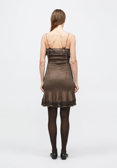 Brun satengkjole med spaghettistropper, rynket livdel og volangkant detaljert med frynser. Brukt med gjennomsiktige svarte strømpebukser og svarte sko.