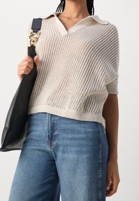 Kremowy, luźny sweter dziany z kołnierzykiem, noszony na niebieskich dżinsach. Trzyma czarną torbę na ramię.