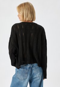 Schwarzer, gestrickter Pullover mit lockerer Passform, der über eine gerippte Textur und ein offenes Strickdesign verfügt. Verkürzte Länge mit rohem Saum. Kombiniert mit blauen Jeans.