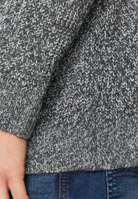 Grauer Strickpullover mit einem strukturierten Muster, das helle und dunkle Fäden enthält. Die Ärmel sind umgeschlagen, und darunter sind Jeans aus Denim sichtbar.