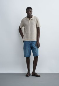 Homme debout contre un mur blanc, portant un polo beige texturé, un short en denim bleu et des sandales en cuir marron.