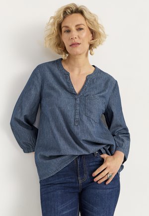 Denimshirt met een V-hals, lange mouwen en een borstzak. Heeft lichte stiksels en een relaxte pasvorm. Draagt bij donkere jeans.