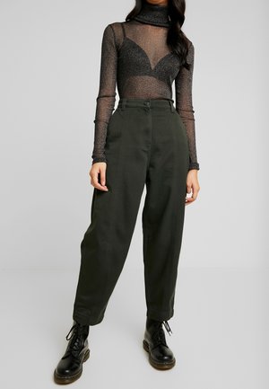 Top negro transparente de manga larga con destellos metálicos, combinado con pantalones de tiro alto en color verde oliva. La modelo lleva botas negras de cordones.