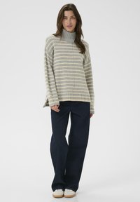 A woman stands wearing a striped, oversized turtleneck sweater and dark wide-leg jeans, showcasing a casual, stylish outfit.