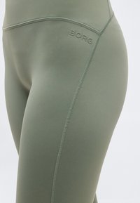 Κοντινό πλάνο ελαστικών κολάν σε ελιά απόχρωση με ραφή στο πλάι και ανάγλυφο λογότυπο "BORG" κοντά στη μέση.