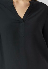 Schwarze Bluse mit V-Ausschnitt und teilweiser Knopfleiste. Sie besticht durch glatten Stoff und einen dezenten Glanz, hat eine lockere Passform und keine sichtbaren Muster.